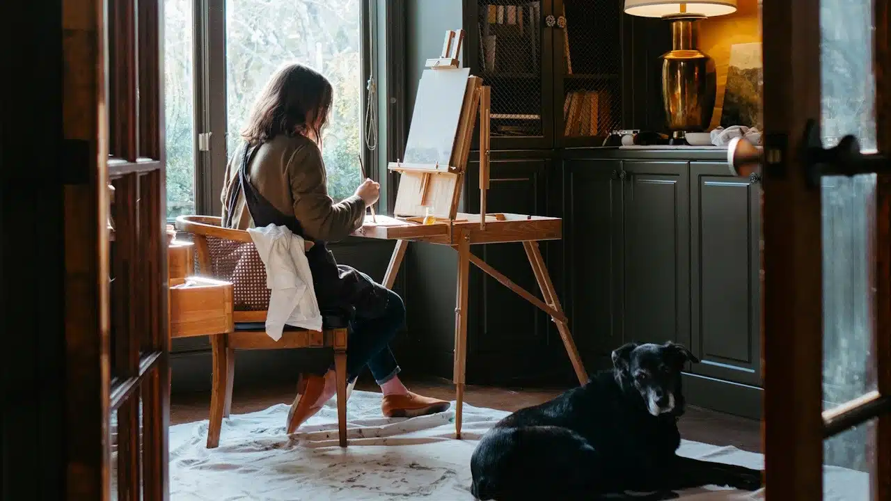 A woman painting on an easel while her dog sits on the ground.