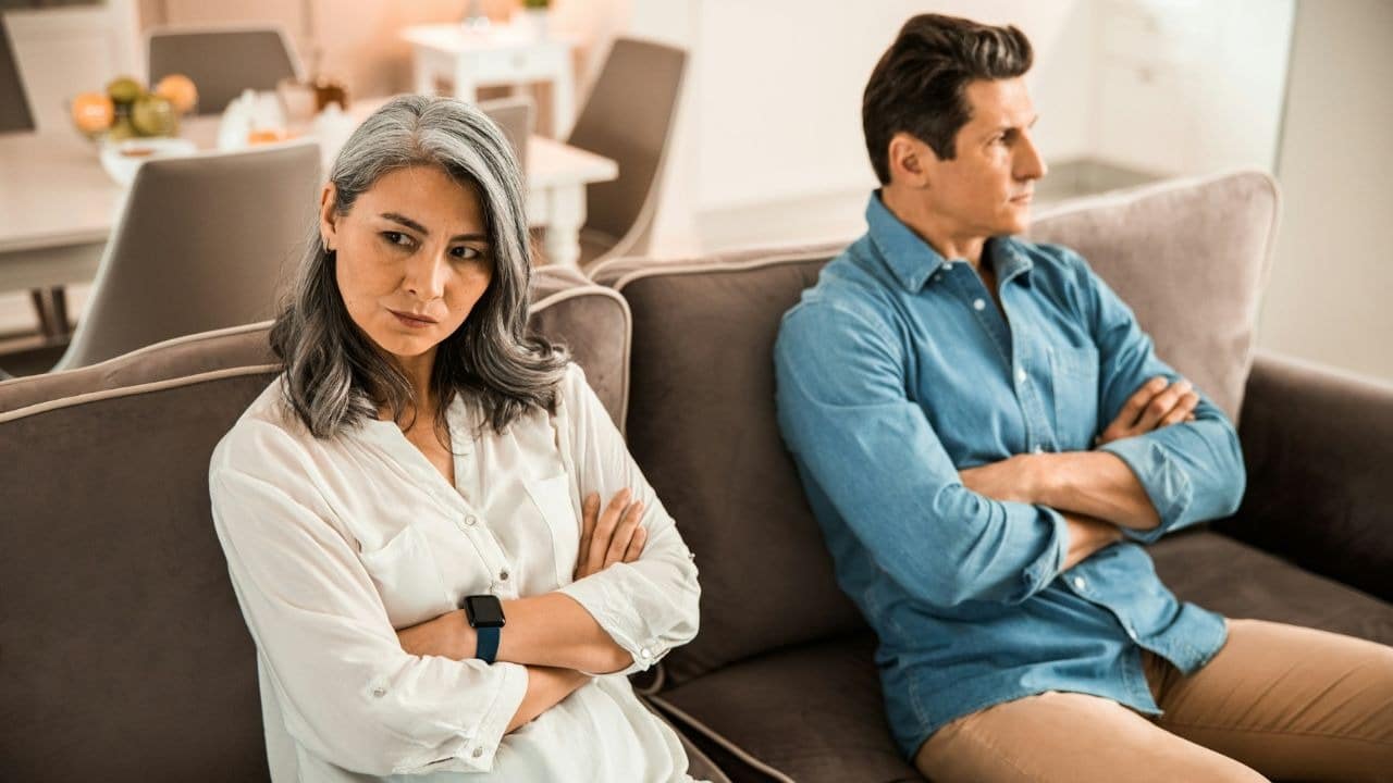 A man and a woman sit on a grey sofa with their arms crossed, looking away.