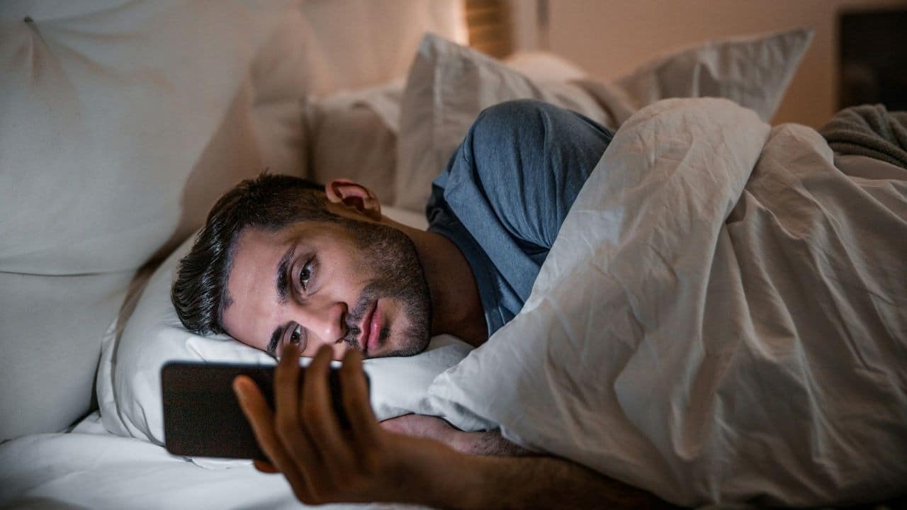 A man lies in a bed under a white blanket while looking at a smartphone.
