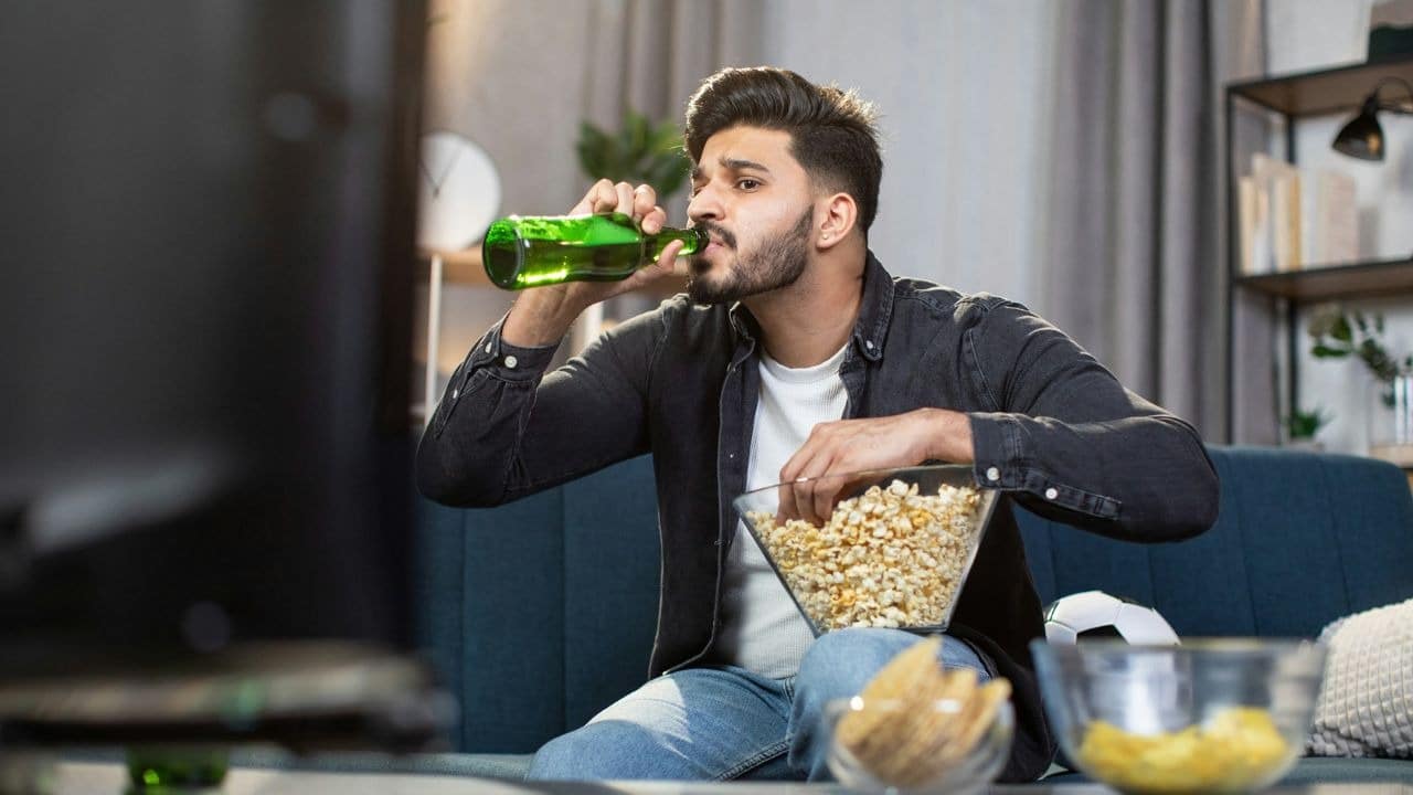 A man sits on a couch drinking from a green bottle while holding a popcorn bowl.
