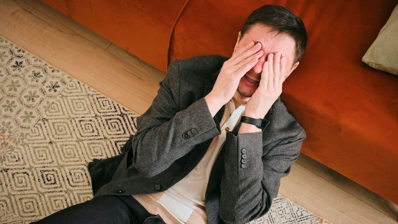 A man in a blazer sits on the floor and covers his face with both hands.