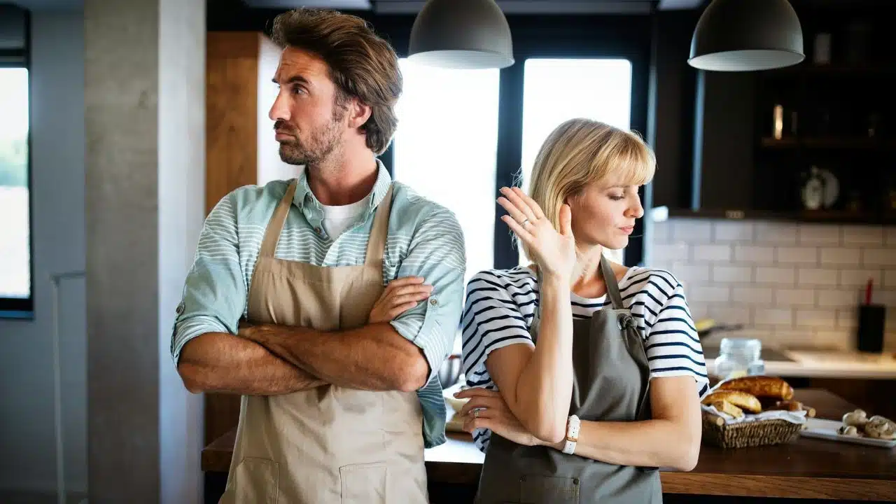 A man and woman in aprons stand back-to-back with arms crossed in a kitchen.