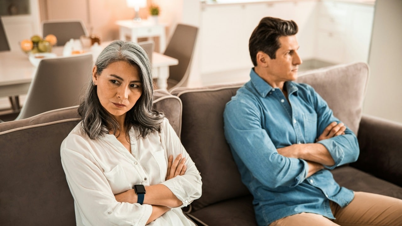 A man and woman sit on a sofa with arms crossed, facing away from each other.