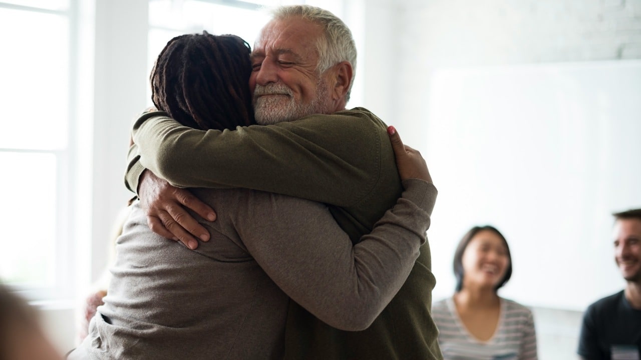 An older man hugging his wife while some people smile in the background.