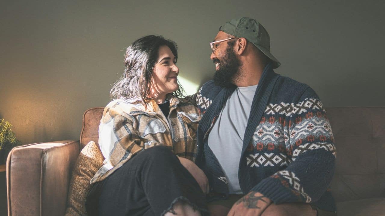 A smiling couple sits close together on a couch, looking at each other.