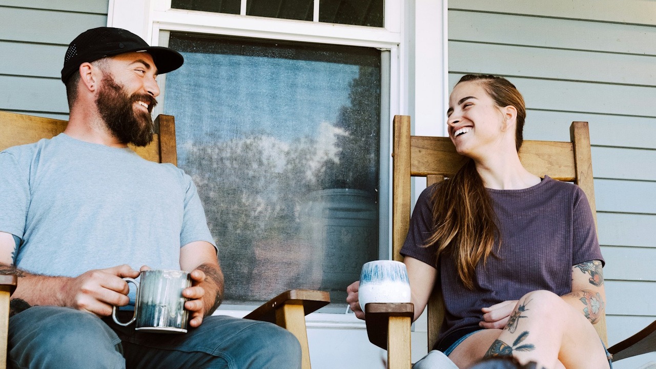 A cheerful couple sitting in wooden rocking chairs on a porch, laughing and holding coffee mugs.
