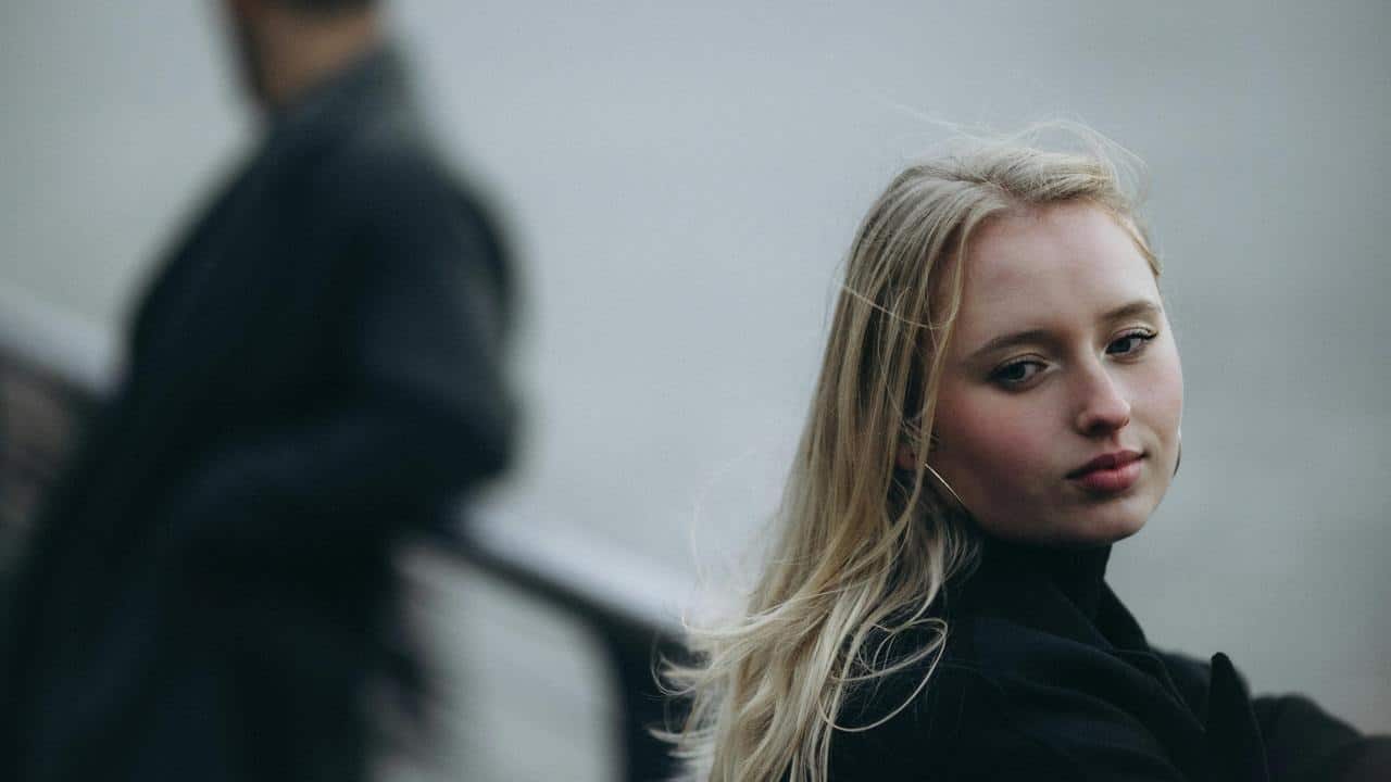 A woman looking away with a man blurred in the background.