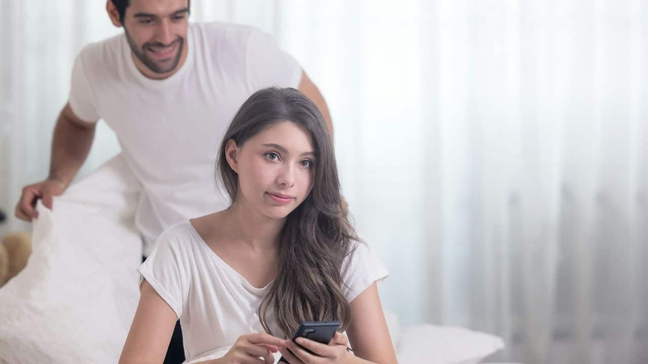 A young woman holds her phone while a man stands smiling in the background.