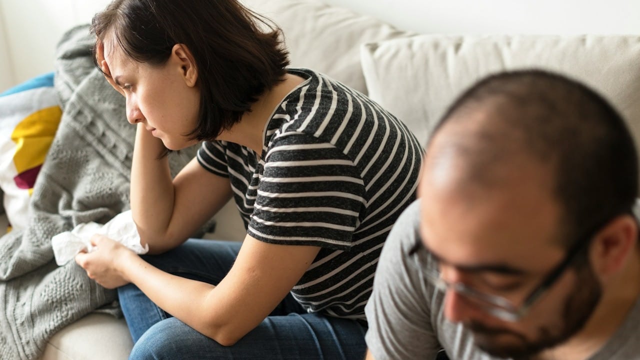 A woman looking upset and a man wearing sunglasses looking down as they sit on a couch.