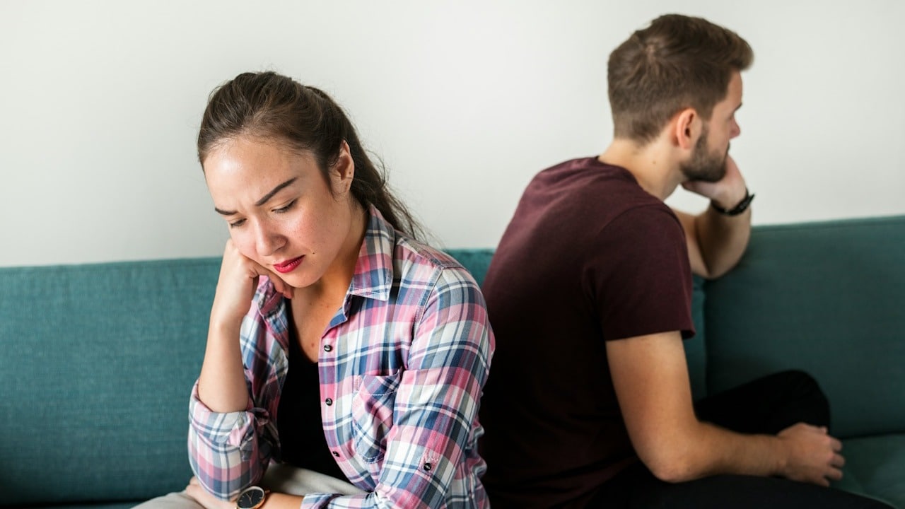 A woman looking sad and sitting with her back to a man who also looks away while they sit on a couch.