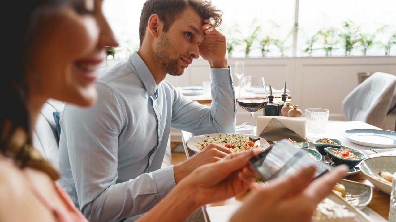 A man with his hand on his head looks at a woman using a phone.