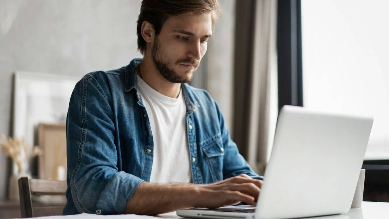 A man with a beard wearing a denim shirt over a white shirt uses a laptop.