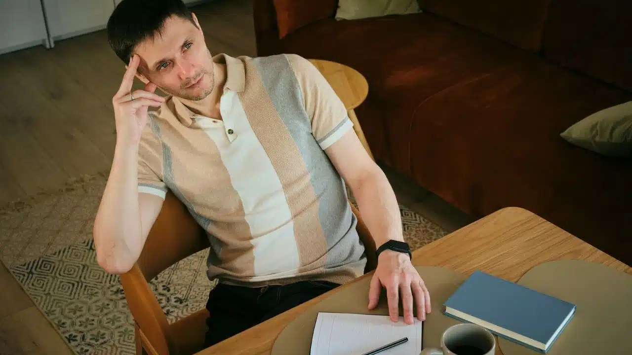A man in a striped polo shirt sits at a wooden table with a notebook.