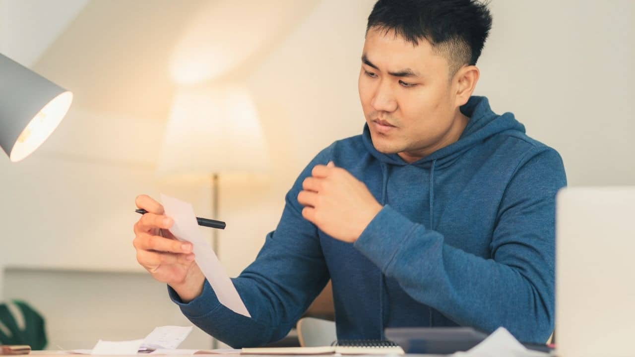 A man in a blue hoodie sits at a desk and looks at a receipt.
