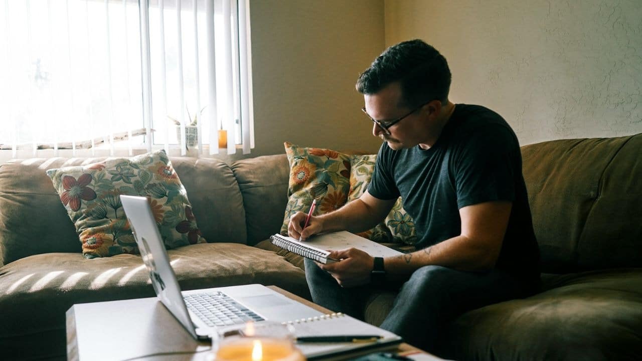 A man with glasses sits on a brown sofa and writes in a spiral notebook.