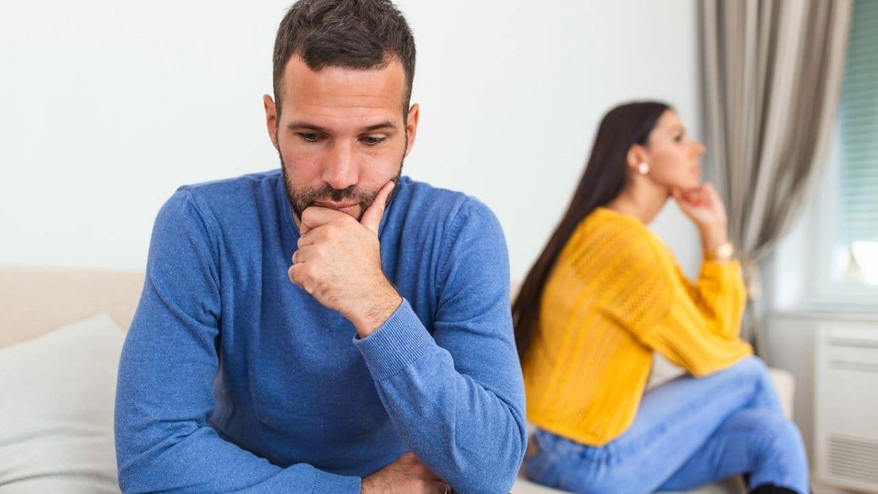 A man in a blue sweater sits on a sofa while a woman looks away.
