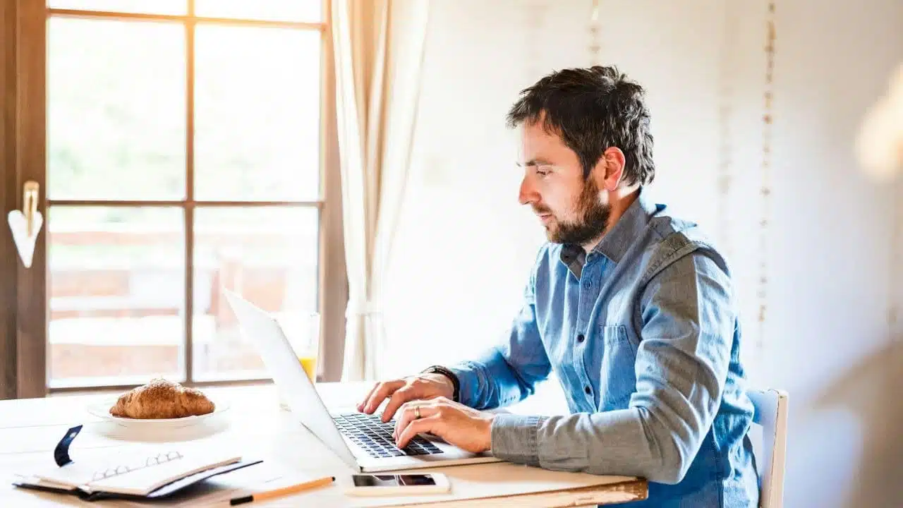 A man in a blue button-down shirt types on a laptop at a white table.