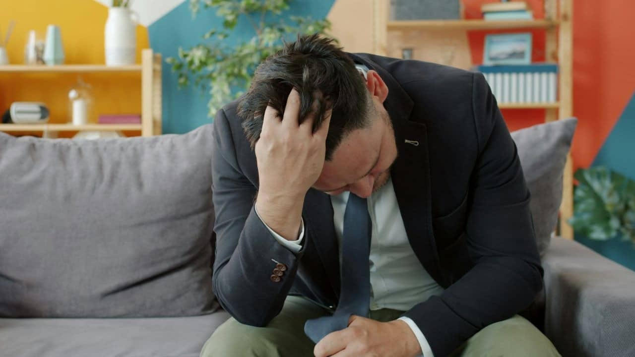 A man in a suit jacket sits on a sofa with his hand on his head.