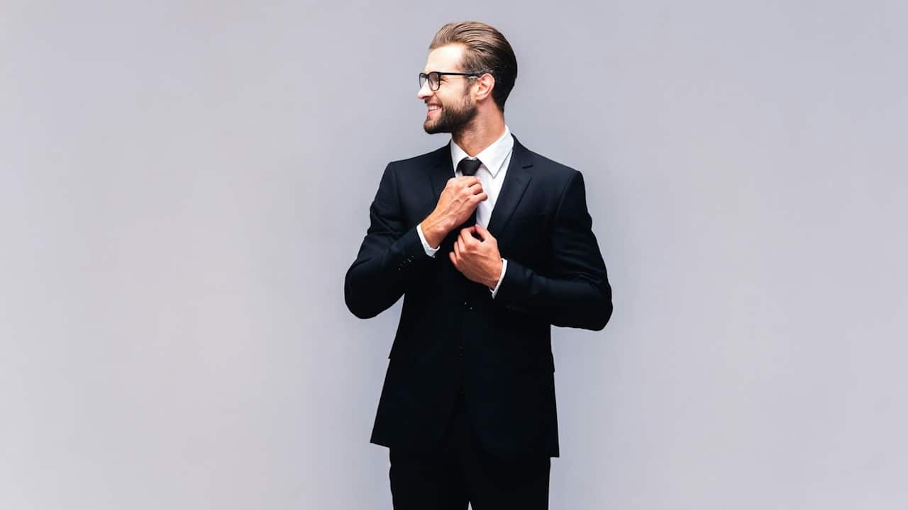 A handsome young man in a full suit adjusting his necktie and smiling.