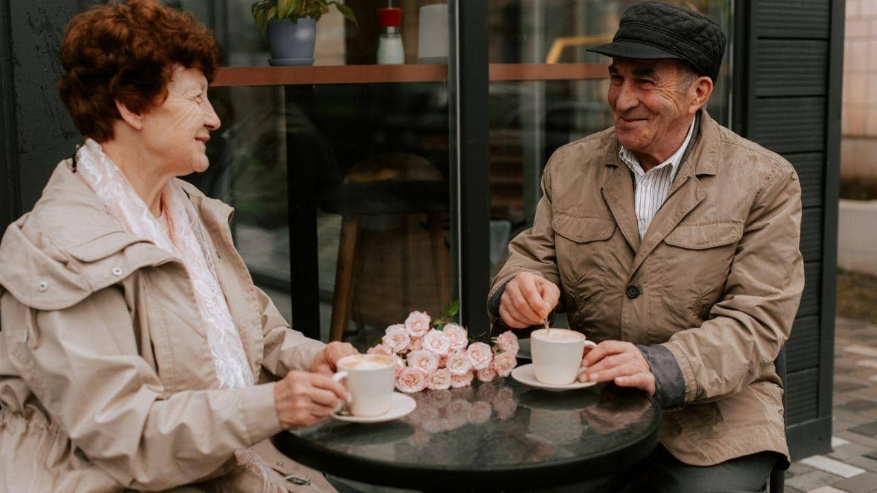 An elderly couple smiles at each other while having coffee at an outdoor table.