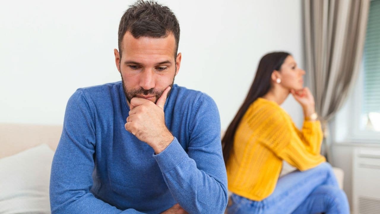 A man sits with his hand on his chin, with a woman sitting out of focus behind him.