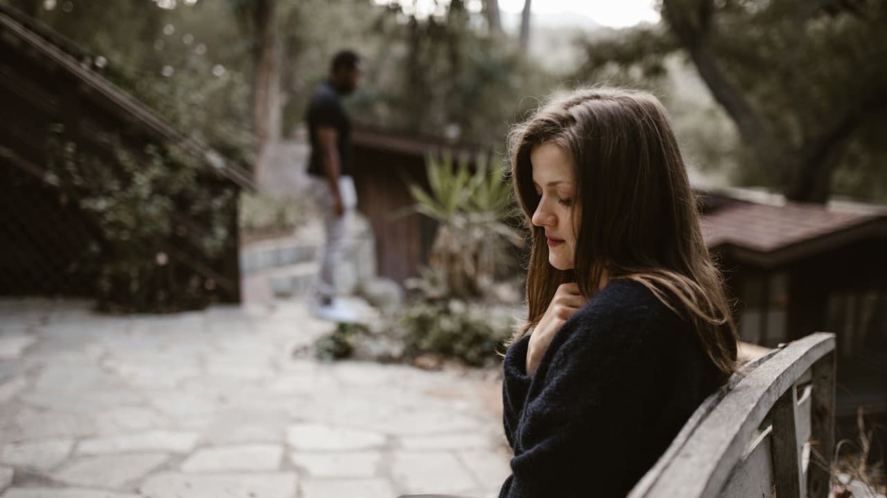 A woman sitting sadly on an outdoor bench with a man standing in the blurry background.