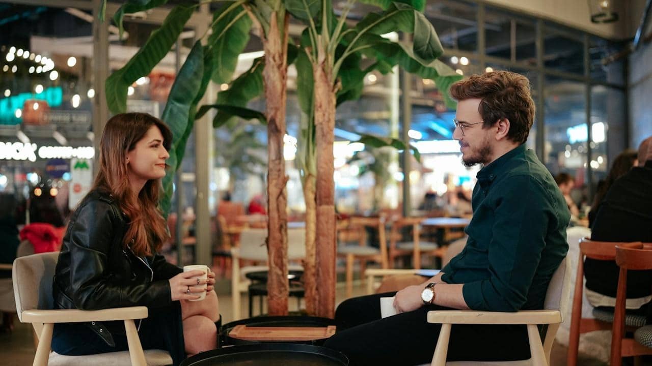 A couple is seated, facing each other in a brightly lit cafe or indoor seating area with plants, having a conversation over coffee.