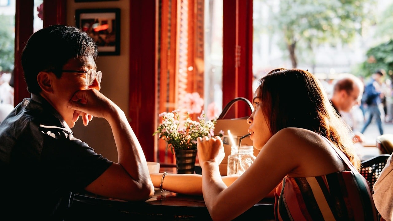 A man wearing a black-collared top sitting on a chair in front of a table and a woman wearing a multicolored top.