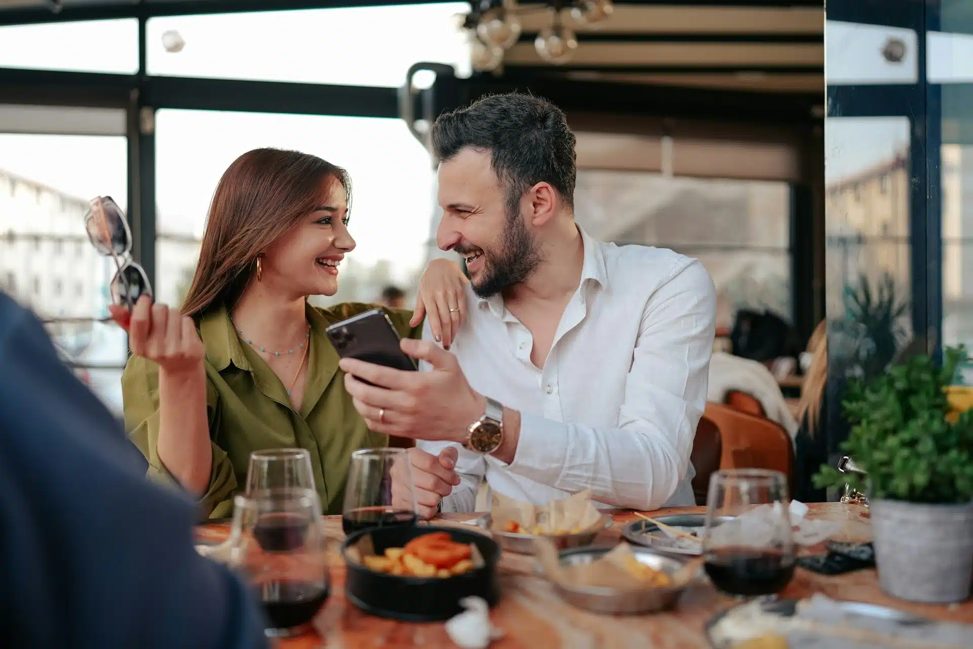A smiling couple sitting at a table while the bearded man shows something to a woman on his phone.