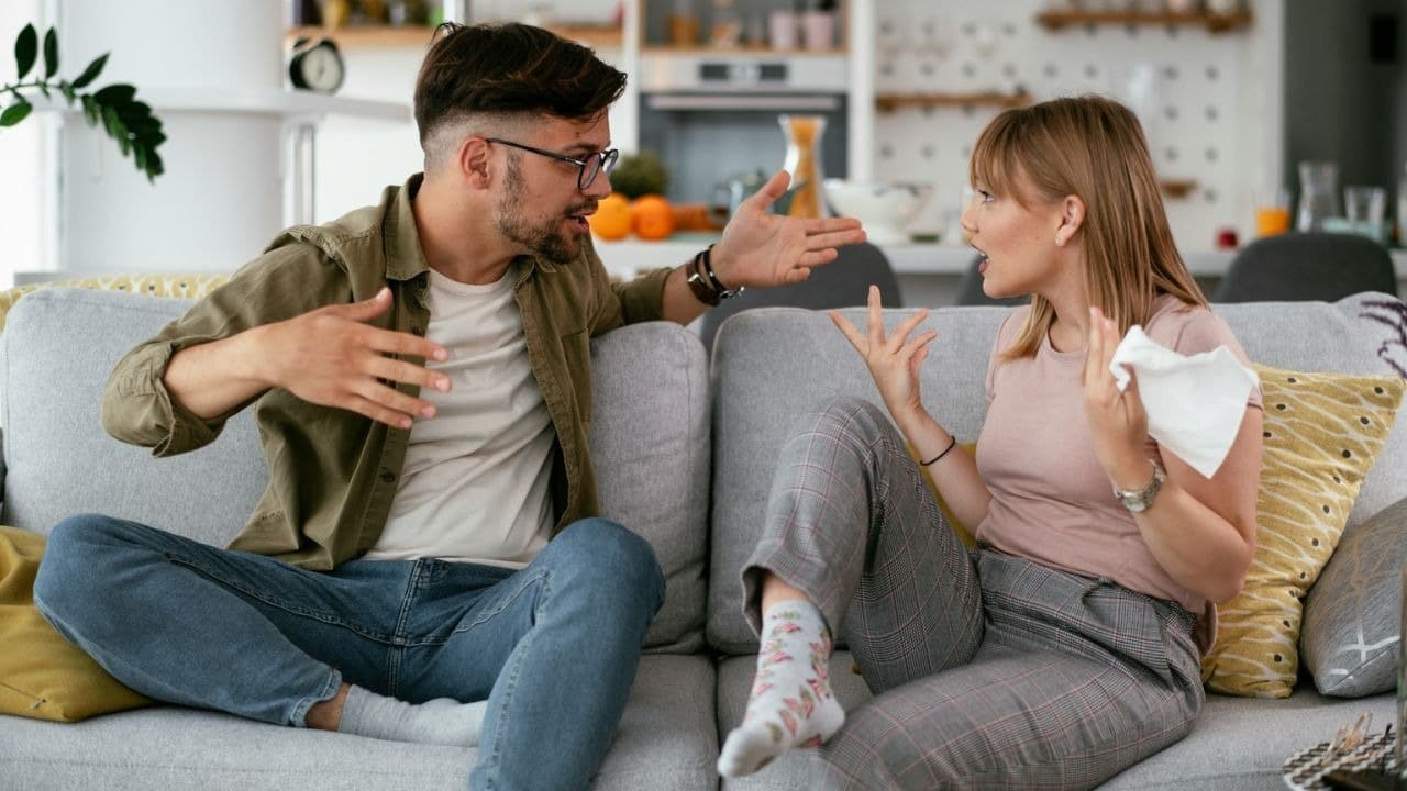 A man and woman are sitting on a couch, gesturing at each other while arguing indoors.