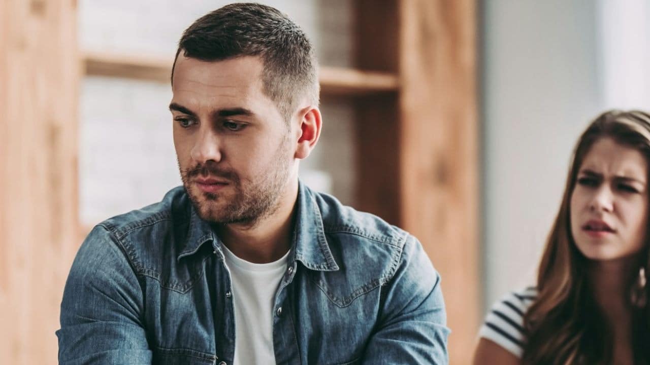 A man with a serious expression is looking down while a frustrated woman is blurry in the background.