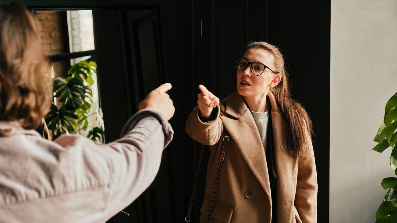 A woman in a tan coat and glasses points angrily at a man