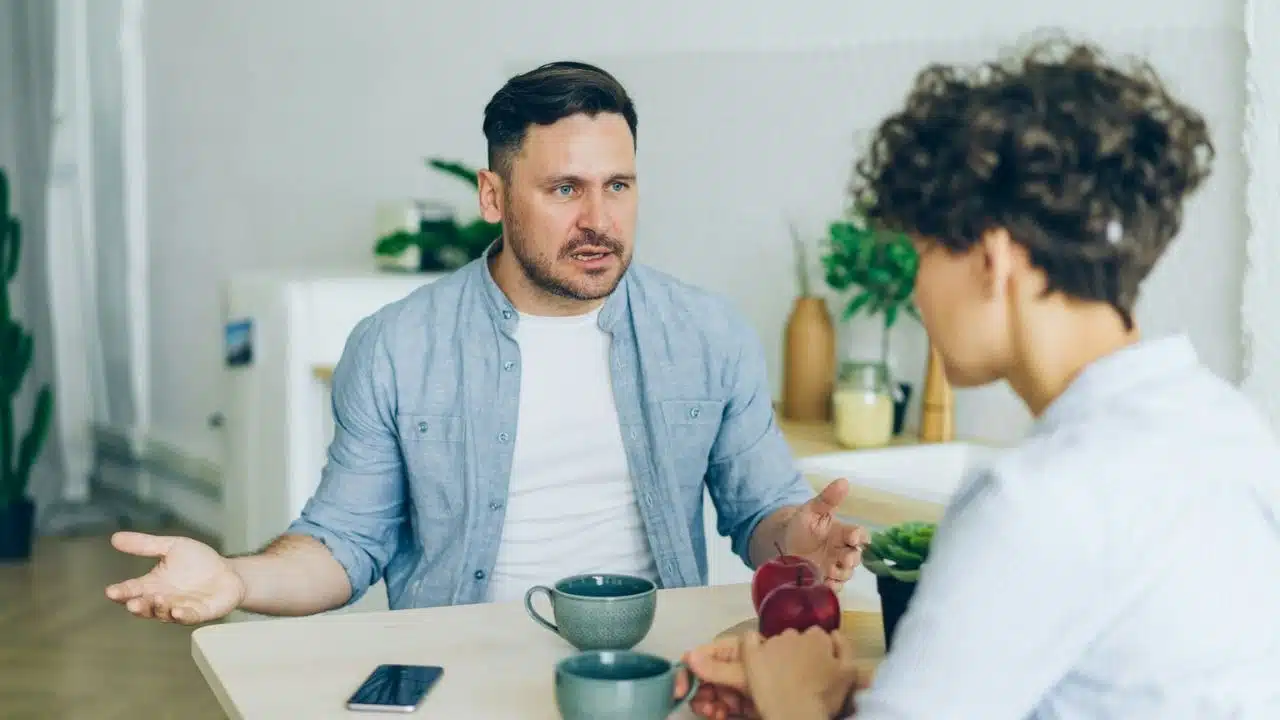 A man is sitting at a table, gesturing with his hands while talking to a woman.