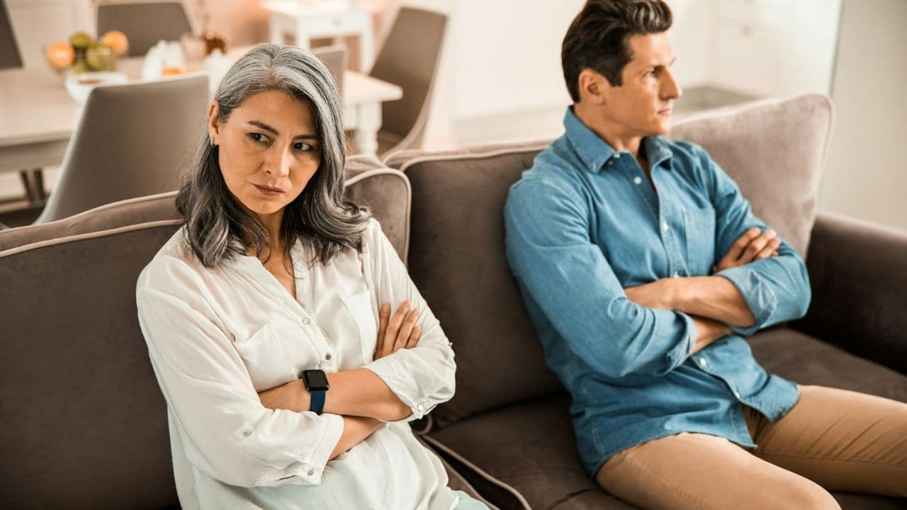 A frustrated, middle-aged couple sits on a couch, facing away from each other with their arms crossed.