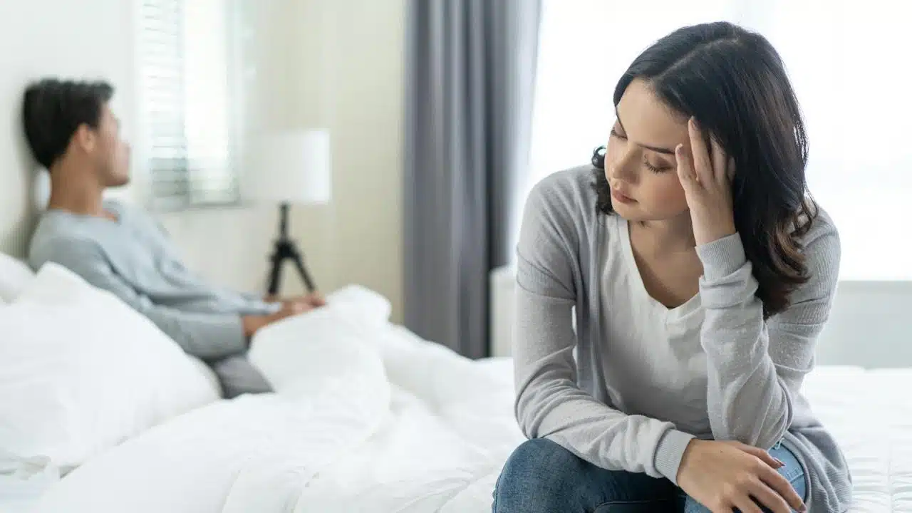 A woman sits on a bed covering her face while a man sits behind her.