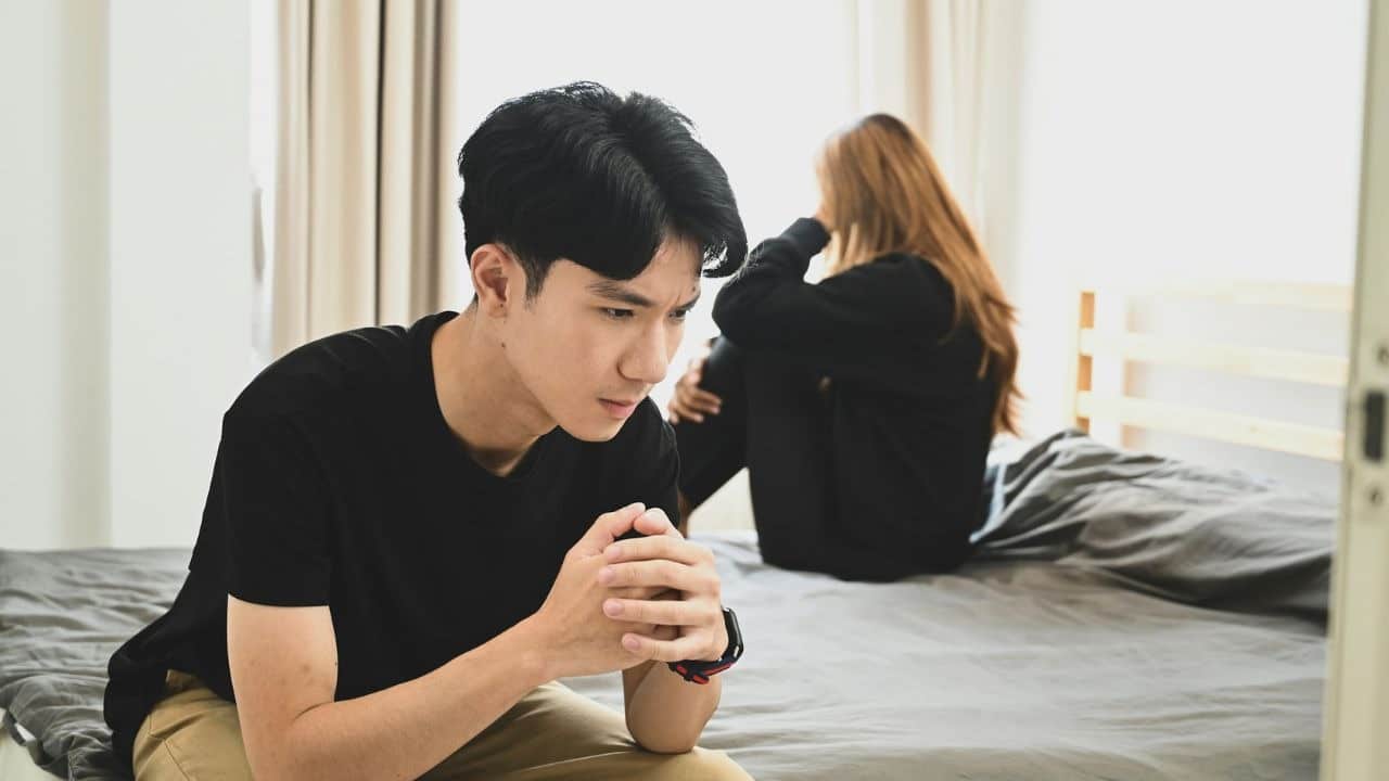 A pensive man sits in the foreground with a sad, crying woman in the background.