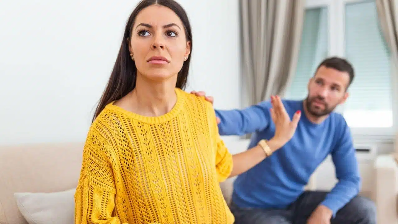 A fed-up woman in a yellow sweater turns away from a man trying to touch her shoulder.