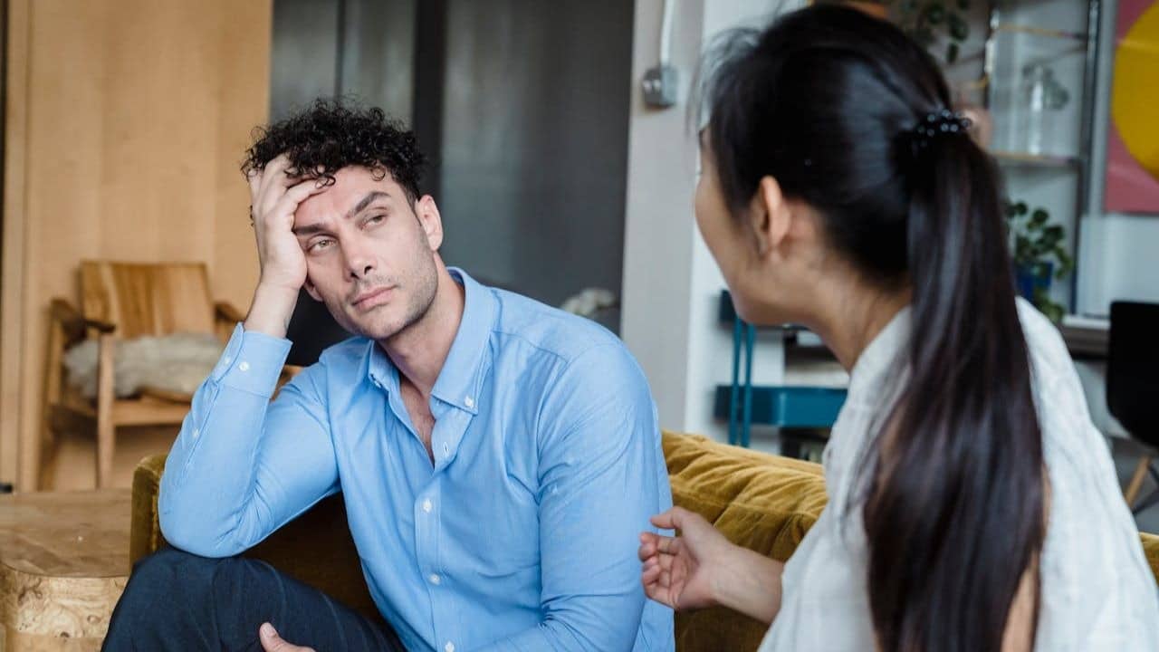 A man looks frustrated, resting his head on his hand while a woman talks to him.