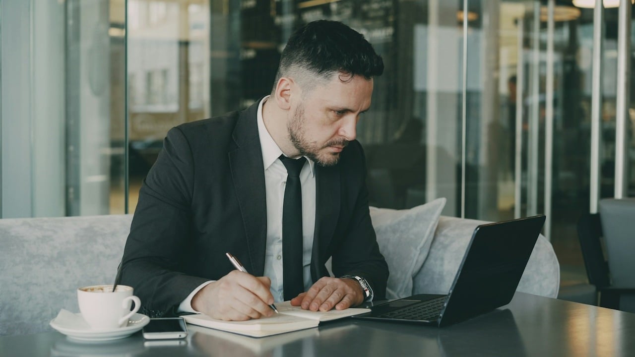 A man writing in his notebook while he looks at something on his laptop.