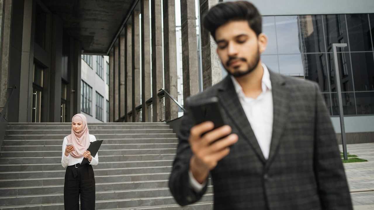 A man in the forefront looking at his phone while a woman wearing a scarf looks at her phone in the background.
