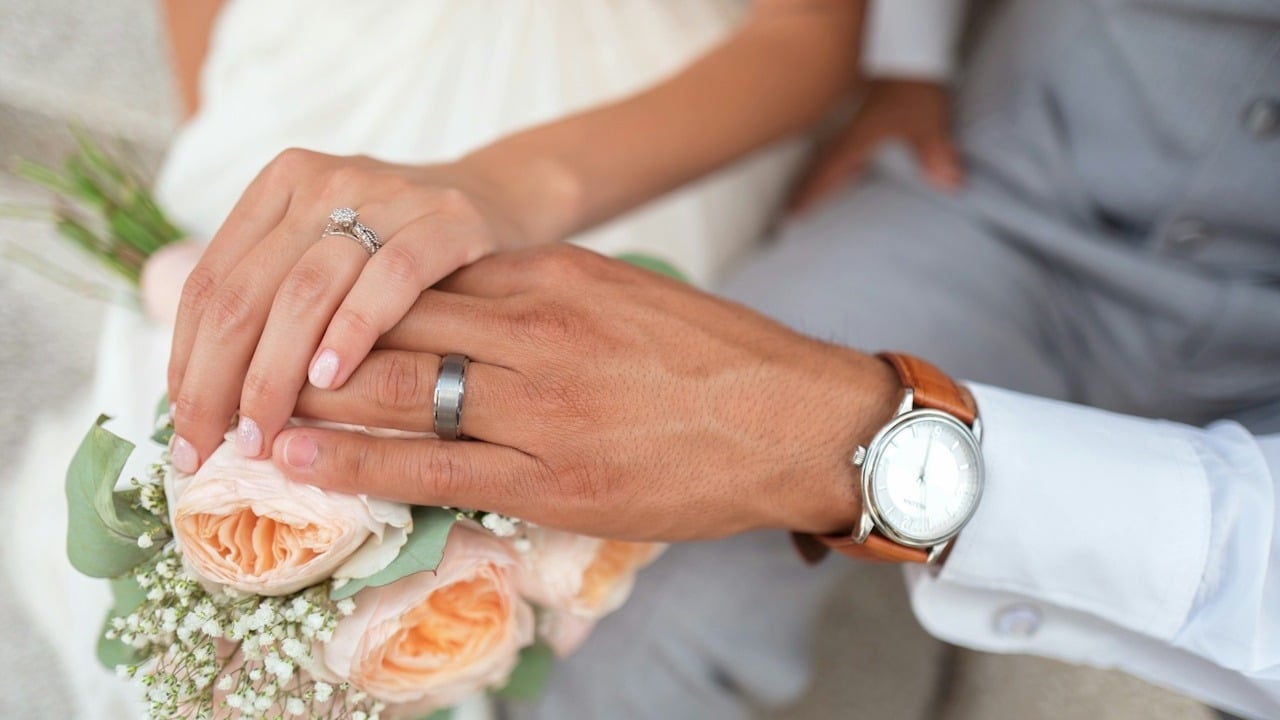 A couple wearing wedding rings touching hands over a bouquet of flowers.