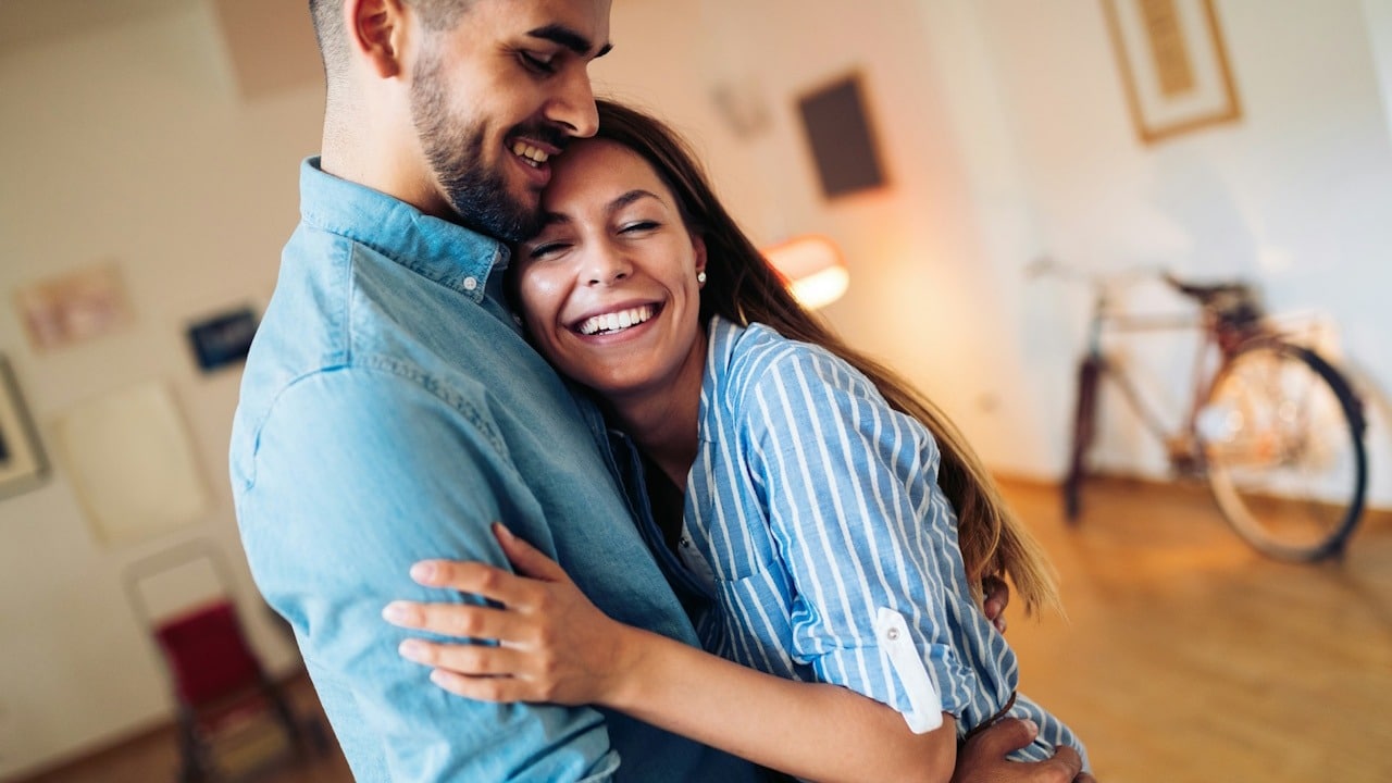 A couple hugging her husband while they are standing indoors.