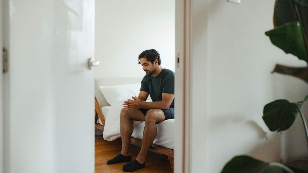 A man sits on the edge of a bed, looking down with his hands clasped.