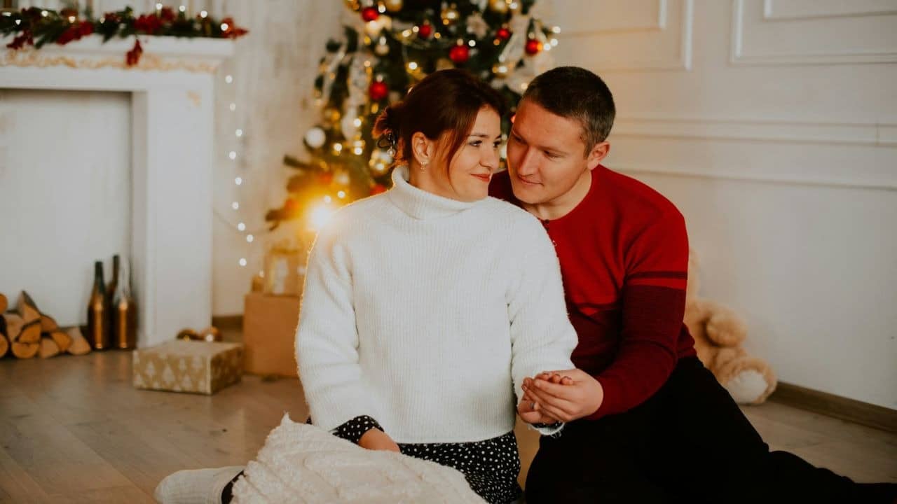 A couple sits on the floor, holding hands, in front of a Christmas tree.