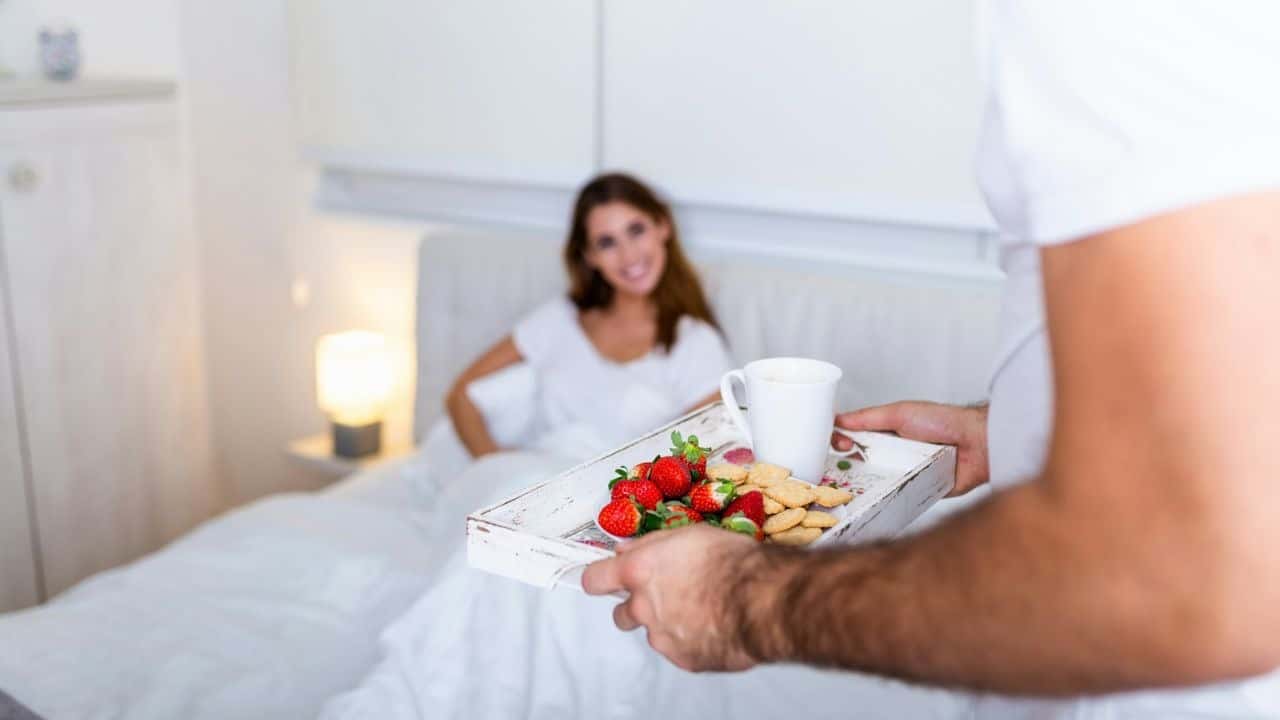 A man with a top bun and glasses looks at a woman sitting on a counter, holding a plate of food.