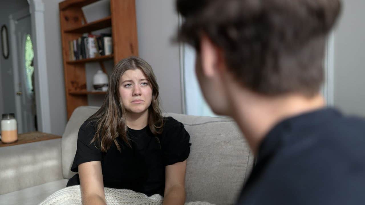 A sad-looking woman sits on a couch, looking at a man whose back is to the camera.