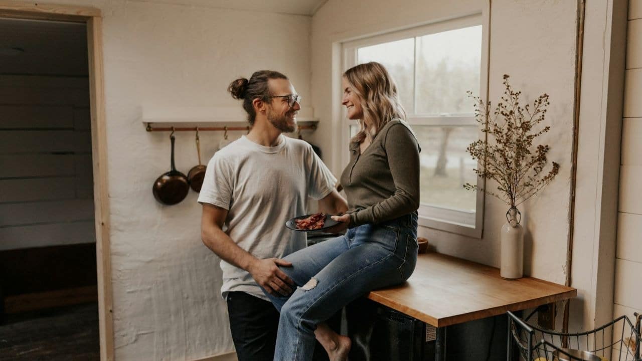 A man with a top bun and glasses looks at a woman sitting on a counter, holding a plate of food.