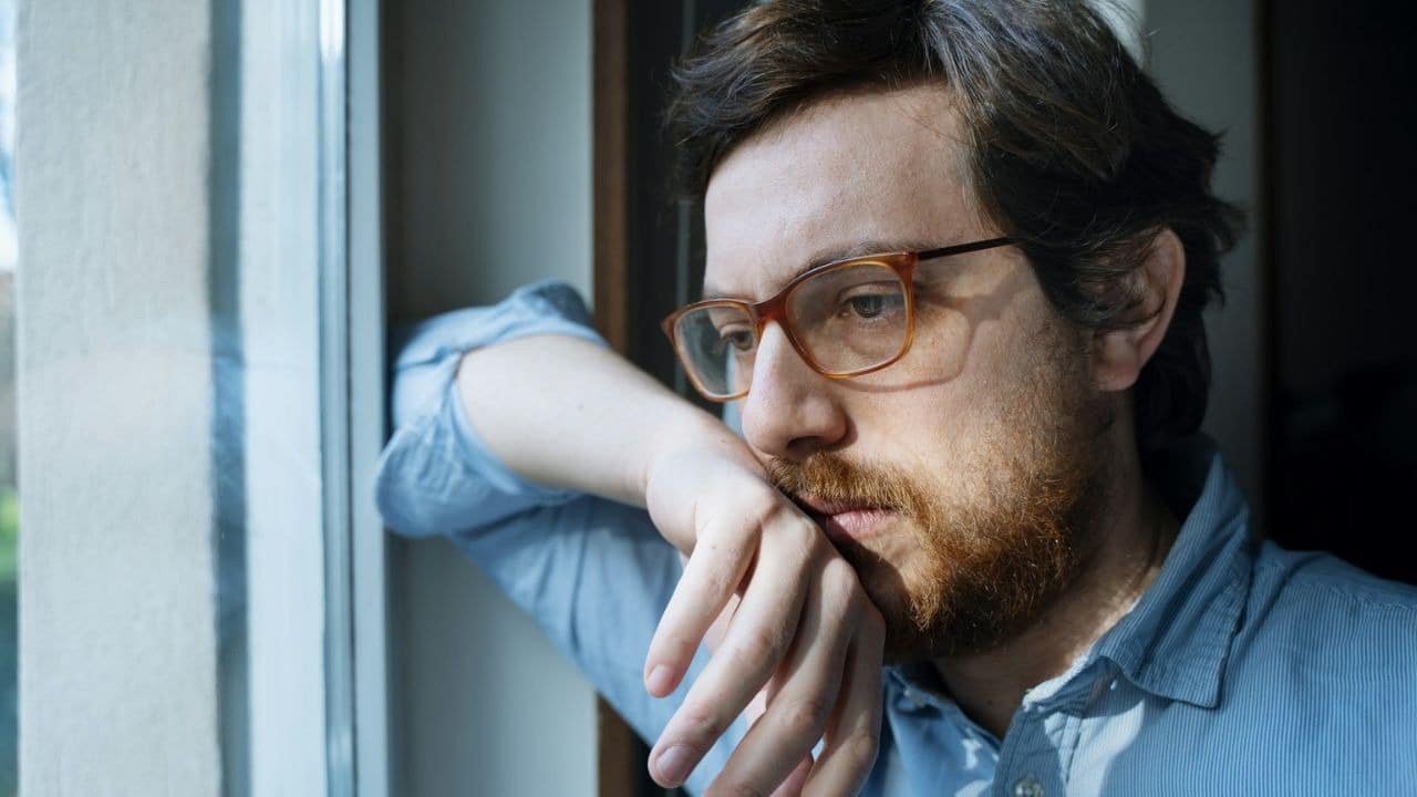 A pensive man with a beard and glasses leans against a window.