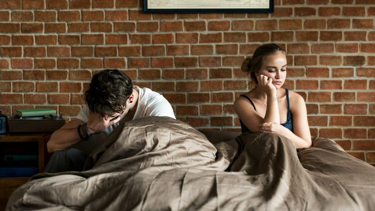 A man and woman sit separately on a bed, both looking distressed.