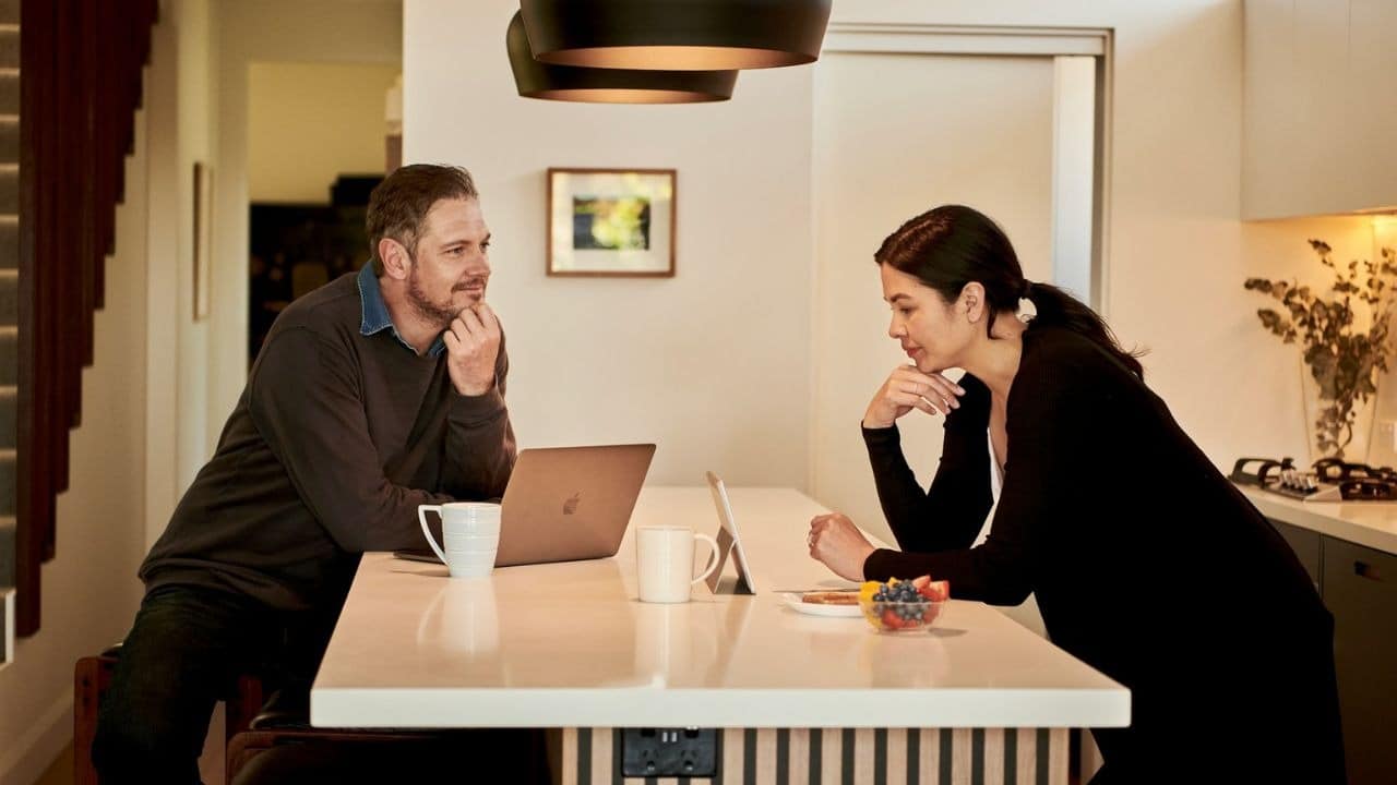 A man on a laptop and a woman with fruit bowls talk across a kitchen island.